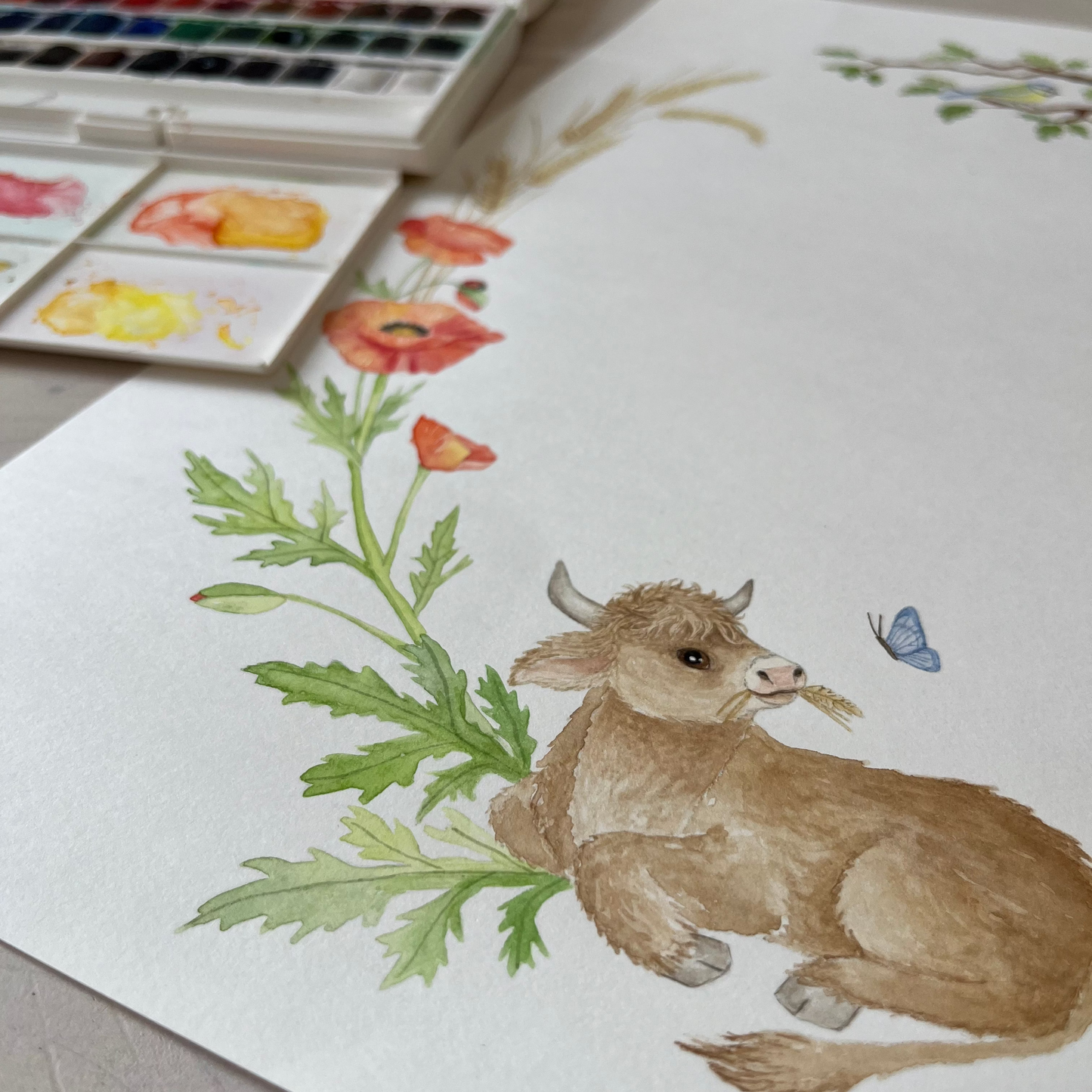 A watercolor painting of a brown calf lying among green leaves and red poppies on white paper, with a watercolor palette visible in the background.
