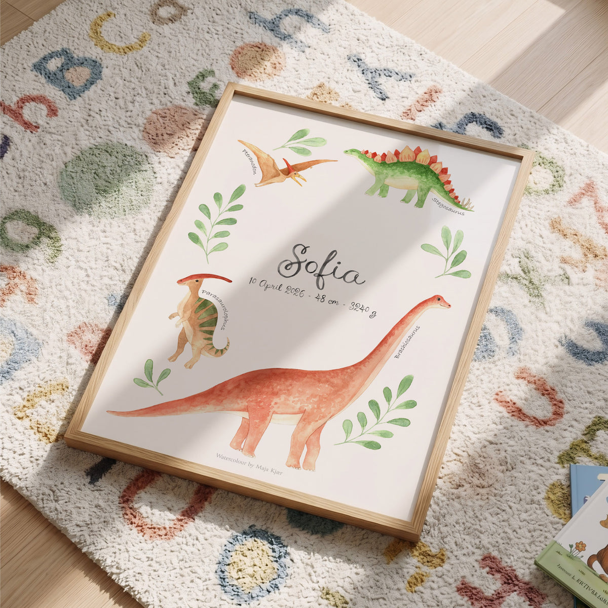 A framed personalized dinosaur art print with watercolor illustrations of dinosaurs and the name 'Sofia' in the center, styled on a colorful alphabet rug on a wooden floor.