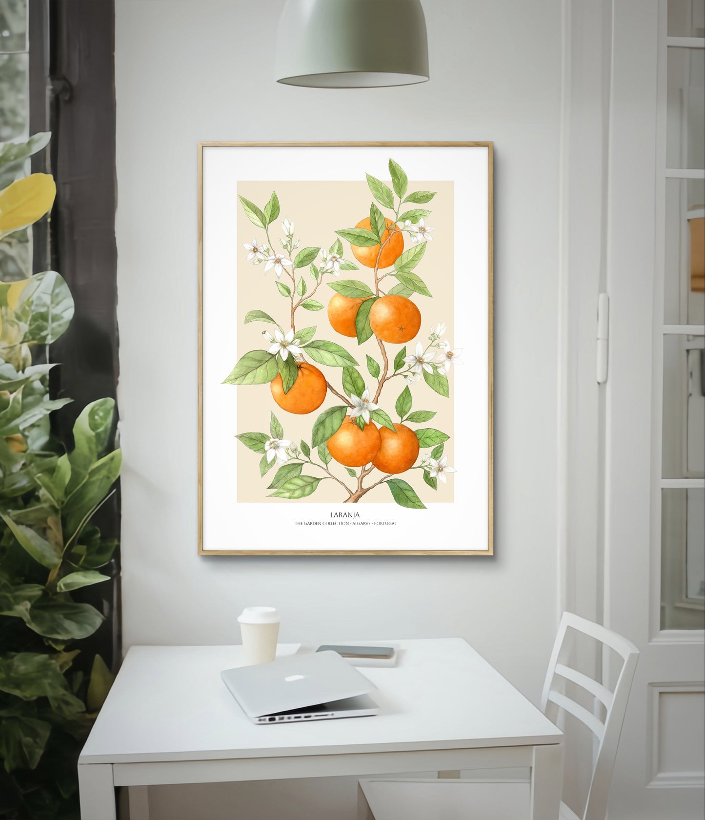 Framed fruit poster with oranges on sand background hanging on above cafe table with laptop and coffee and a violin fig potted plant to the side