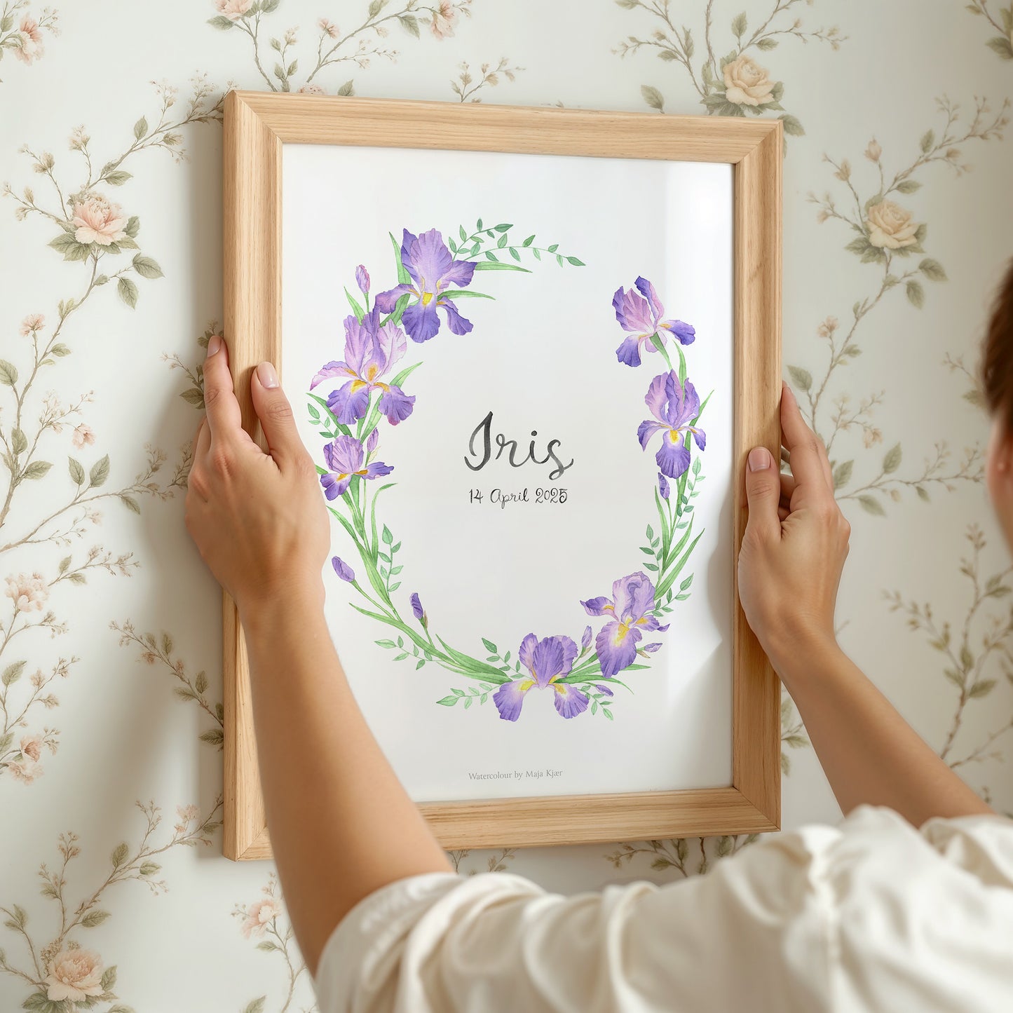 Person holding a framed print with purple flowers and 'Iris' text against a floral wallpaper background.