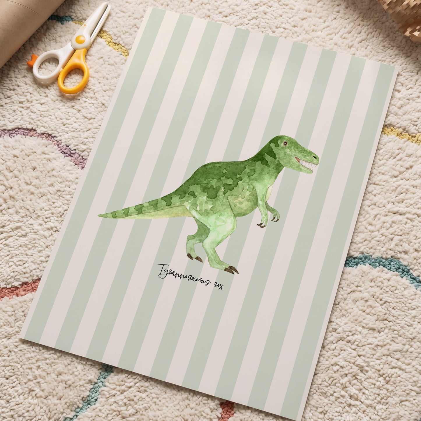 A children's art print featuring a green watercolor T-rex dinosaur on a mint green and white striped background, styled on a textured beige carpet with craft scissors and a cardboard tube nearby.