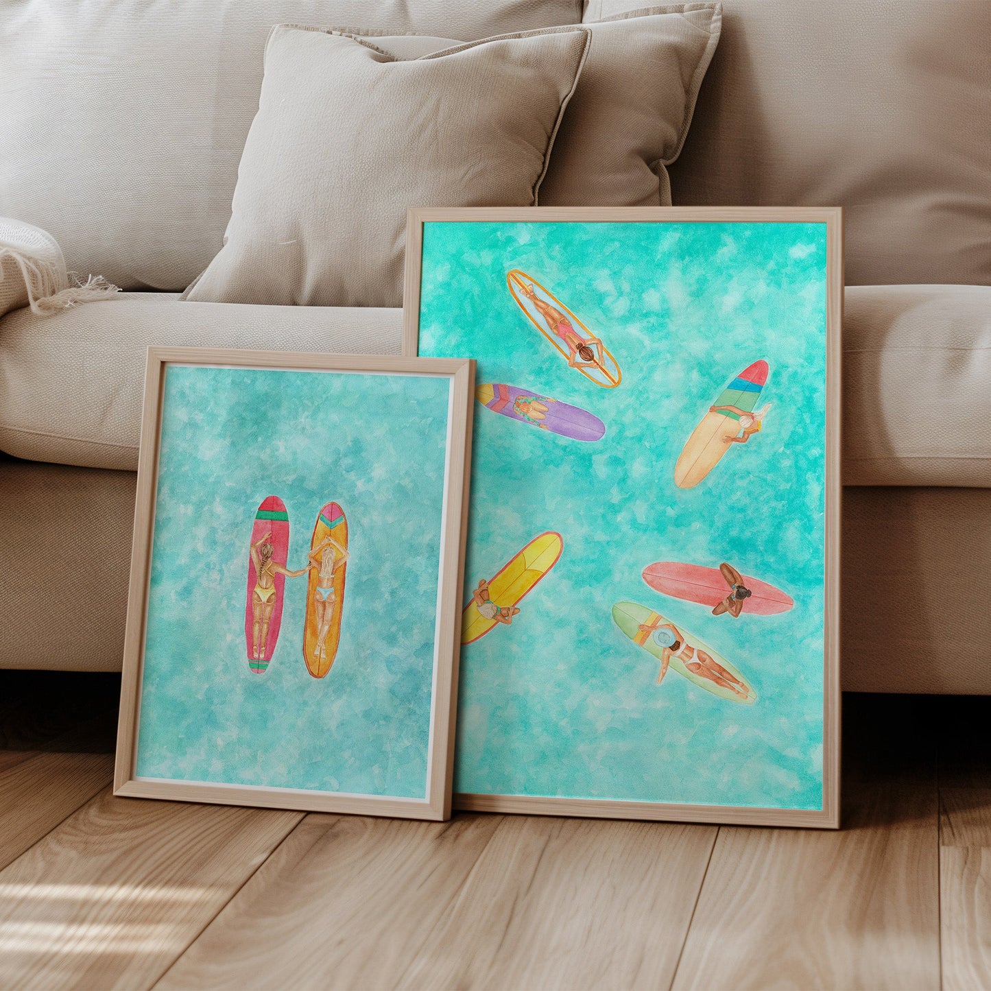 Two framed artworks of colorful surfboards on a wooden floor with a beige sofa in the background.