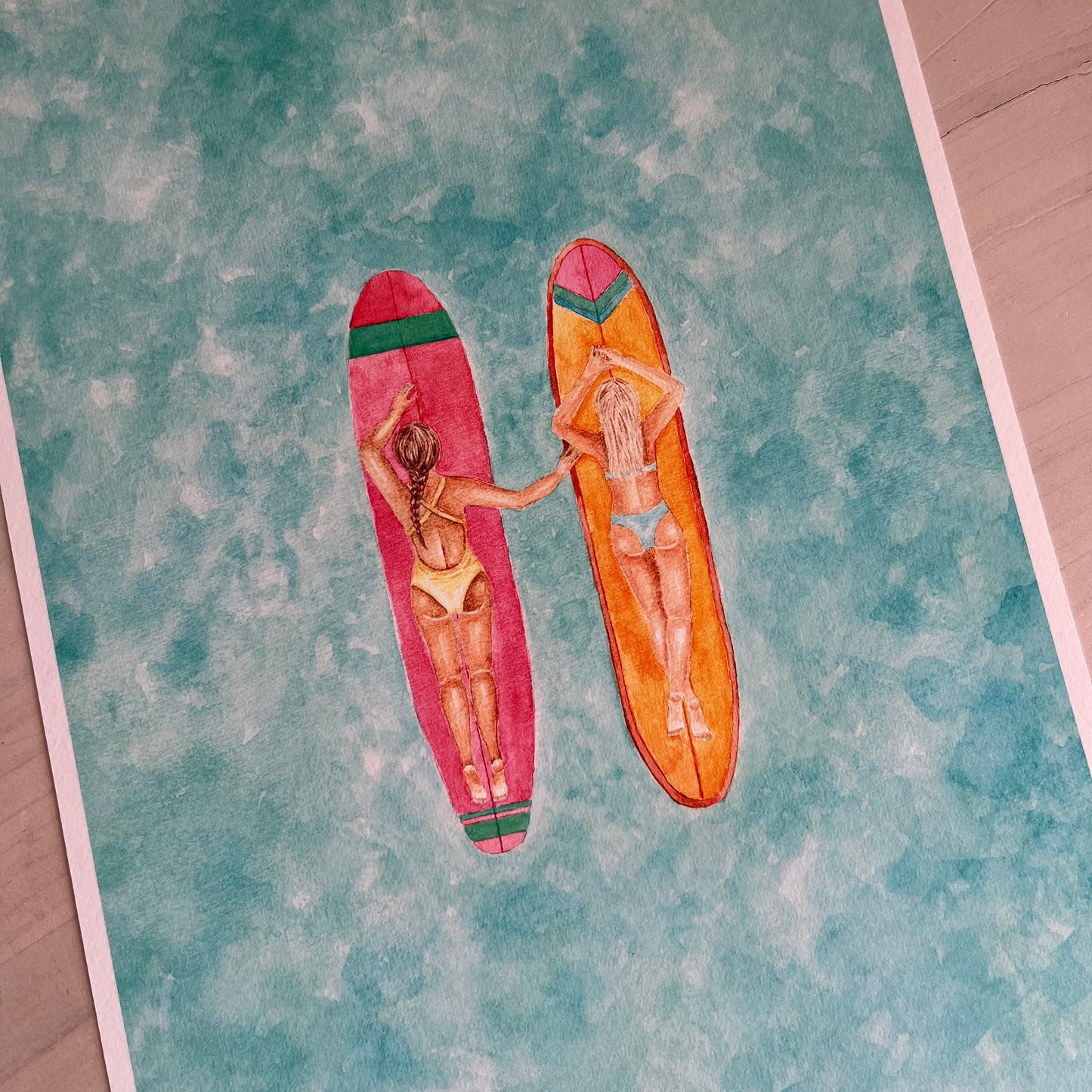 Two women lying on surfboards with a colorful pattern on a textured surface.