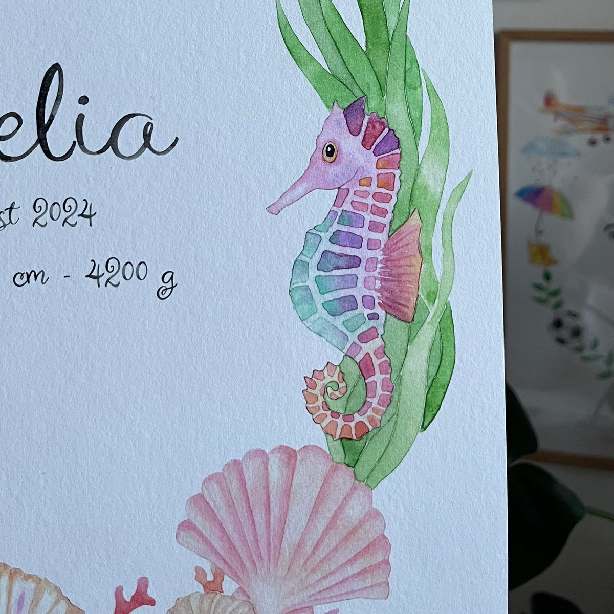 A close-up of a white baby shower card featuring watercolor illustrations of a seahorse, starfish, seashells, and green seaweed, with partial black script text on the left.