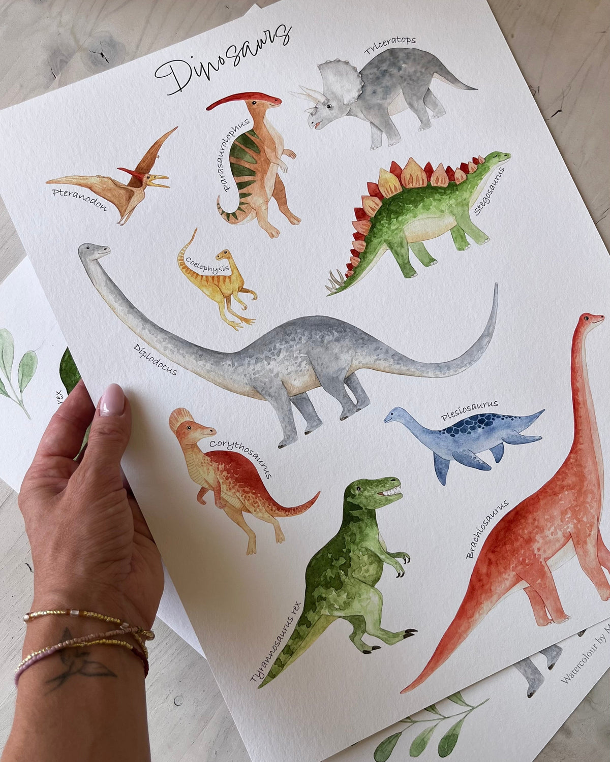 A hand holds a printed watercolor dinosaur poster over another matching print on a light wood surface. The poster features colorful illustrated dinosaurs including a long-necked sauropod, a T-rex, a Stegosaurus, and a Triceratops, with the title 'Dinosaur' at the top.