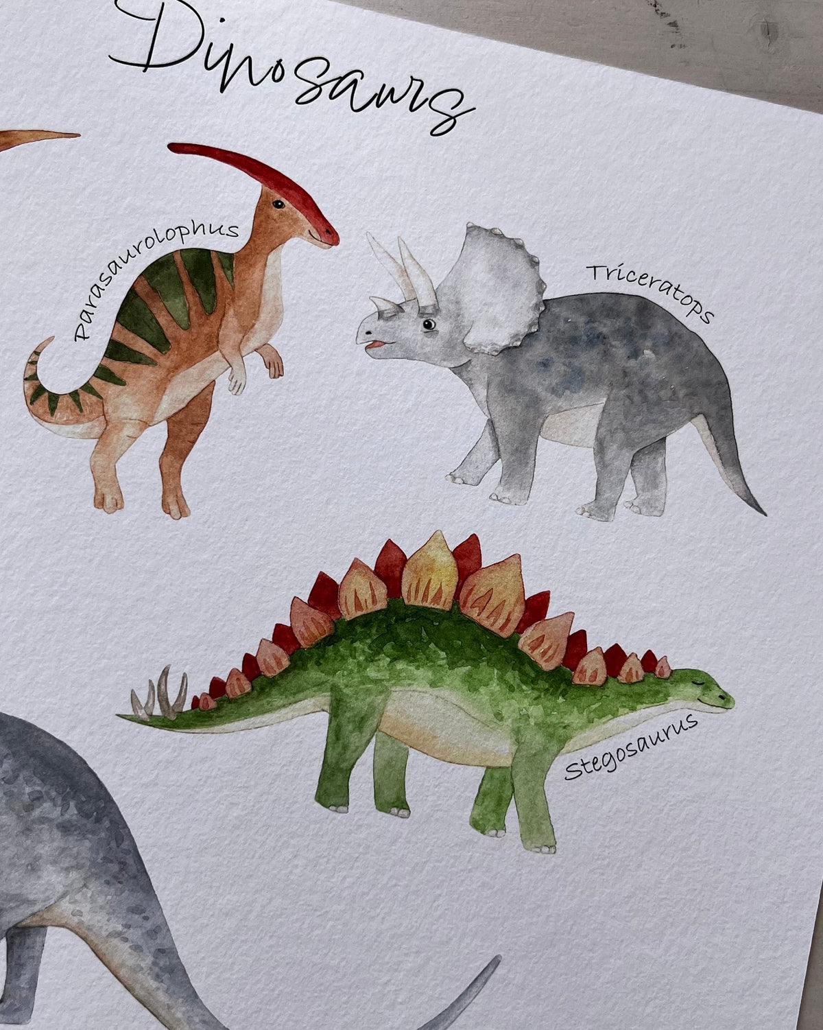 A watercolor dinosaur art print featuring labeled illustrations of dinosaurs including Stegosaurus, Triceratops, and others, photographed on a light wood surface.