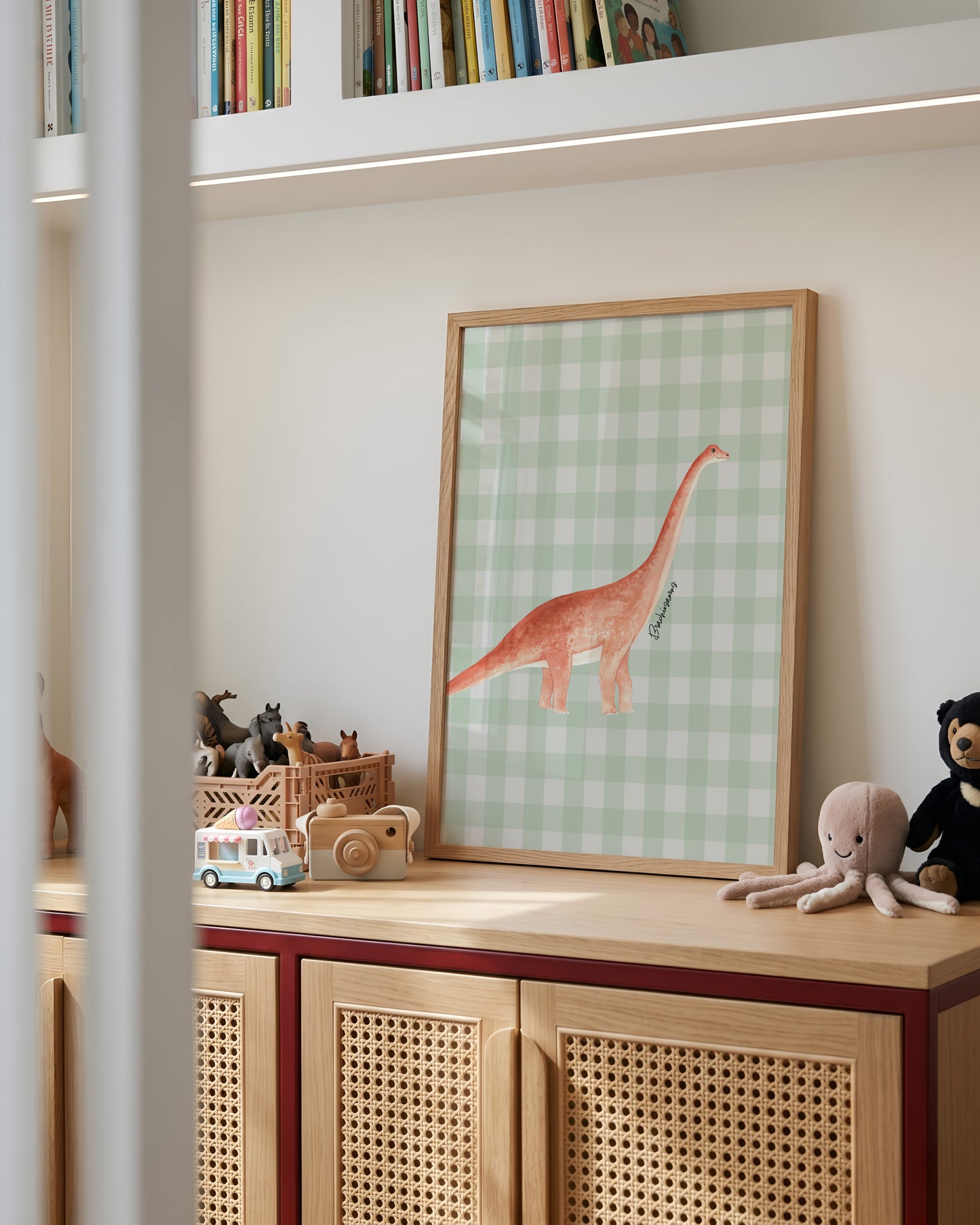 A framed art print featuring a pinkish-orange long-neck dinosaur on a light green gingham background, styled on a wooden cabinet in a child's room.