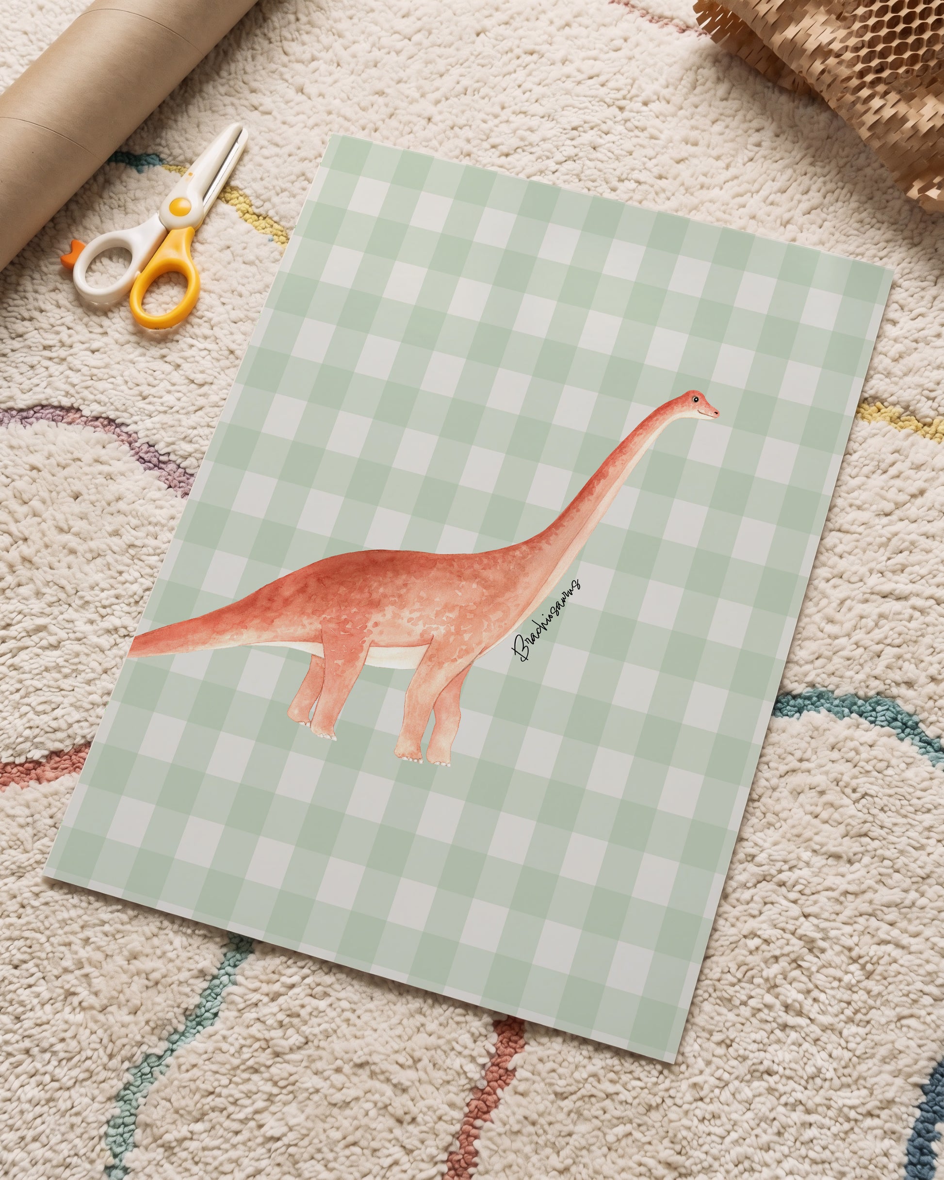 A square art print featuring a watercolor illustration of a long-necked dinosaur on a light green and white gingham checkered background, styled on a textured cream rug with craft scissors and a cardboard tube nearby.