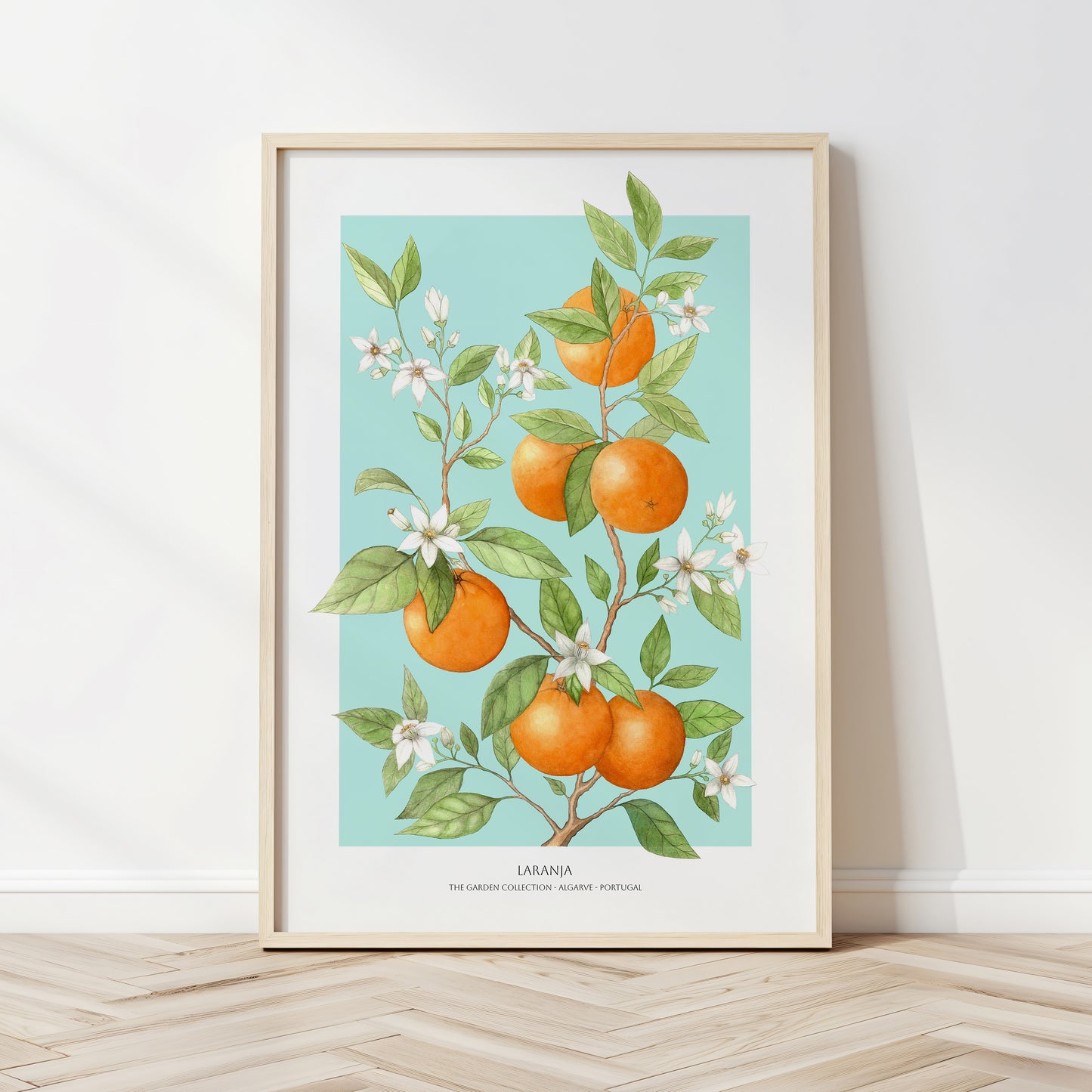 Framed artwork of oranges and flowers on a light blue background, leaning against a white wall.