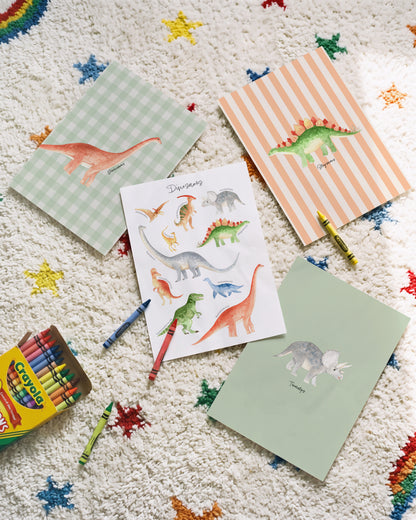 A styled flat-lay of children's dinosaur-themed stationery on a textured white rug with colorful star patterns. The set includes a box of Crayola crayons, a striped dinosaur notepad, a gingham dinosaur notepad, a dinosaur sticker sheet, and a plain green card, with a few crayons placed around them.