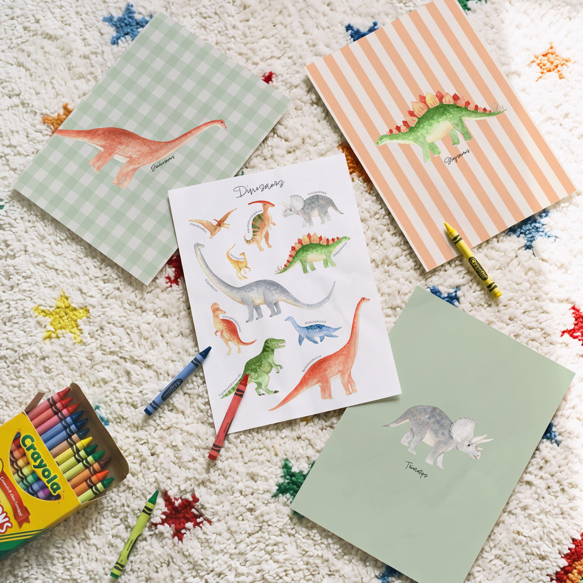 A styled flat-lay of children's dinosaur-themed stationery on a textured white rug with colorful star patterns. The set includes a box of Crayola crayons, a striped dinosaur notepad, a gingham dinosaur notepad, a dinosaur sticker sheet, and a plain green card, with a few crayons placed around them.