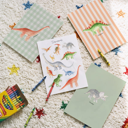 A styled flat-lay of children's dinosaur-themed stationery on a textured white rug with colorful star patterns. The set includes a box of Crayola crayons, a striped dinosaur notepad, a gingham dinosaur notepad, a dinosaur sticker sheet, and a plain green card, with a few crayons placed around them.