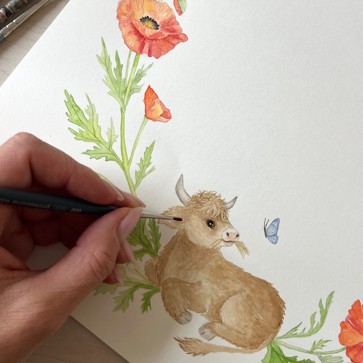 A hand holds a paintbrush over a watercolor painting of a small tan calf surrounded by red poppies, green leaves, and wheat on white paper.