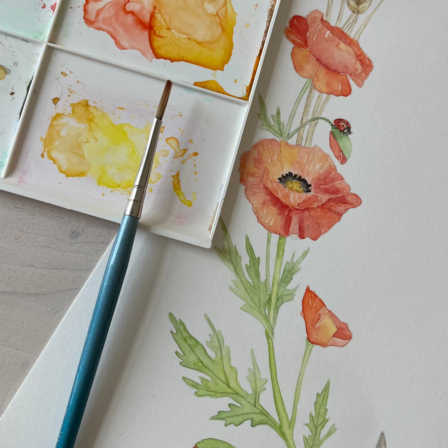 A close-up of a watercolor painting in progress, showing a paint palette with orange, yellow, and pink washes, a fine-tip paintbrush, and a white sheet of paper with a floral and animal illustration.