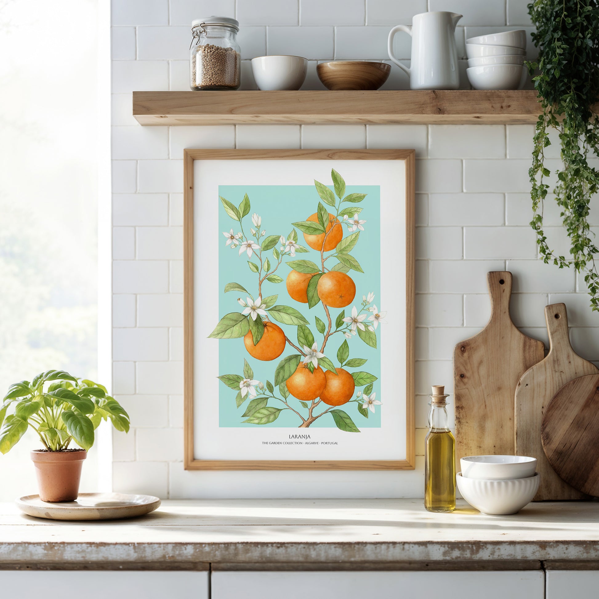 Framed botanical print of oranges on a kitchen counter with wooden cutting boards and a plant.