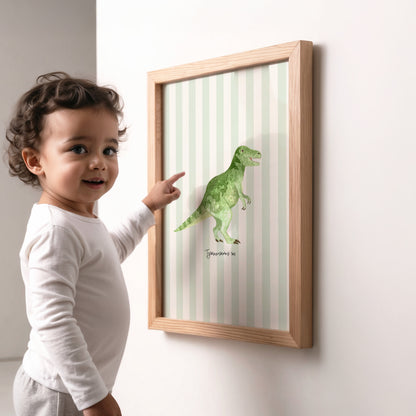 A toddler in a white long-sleeve shirt points at a framed dinosaur art print hanging on a white wall. The print features a green watercolor T-rex on a pale green and white striped background, enclosed in a light natural wood frame.