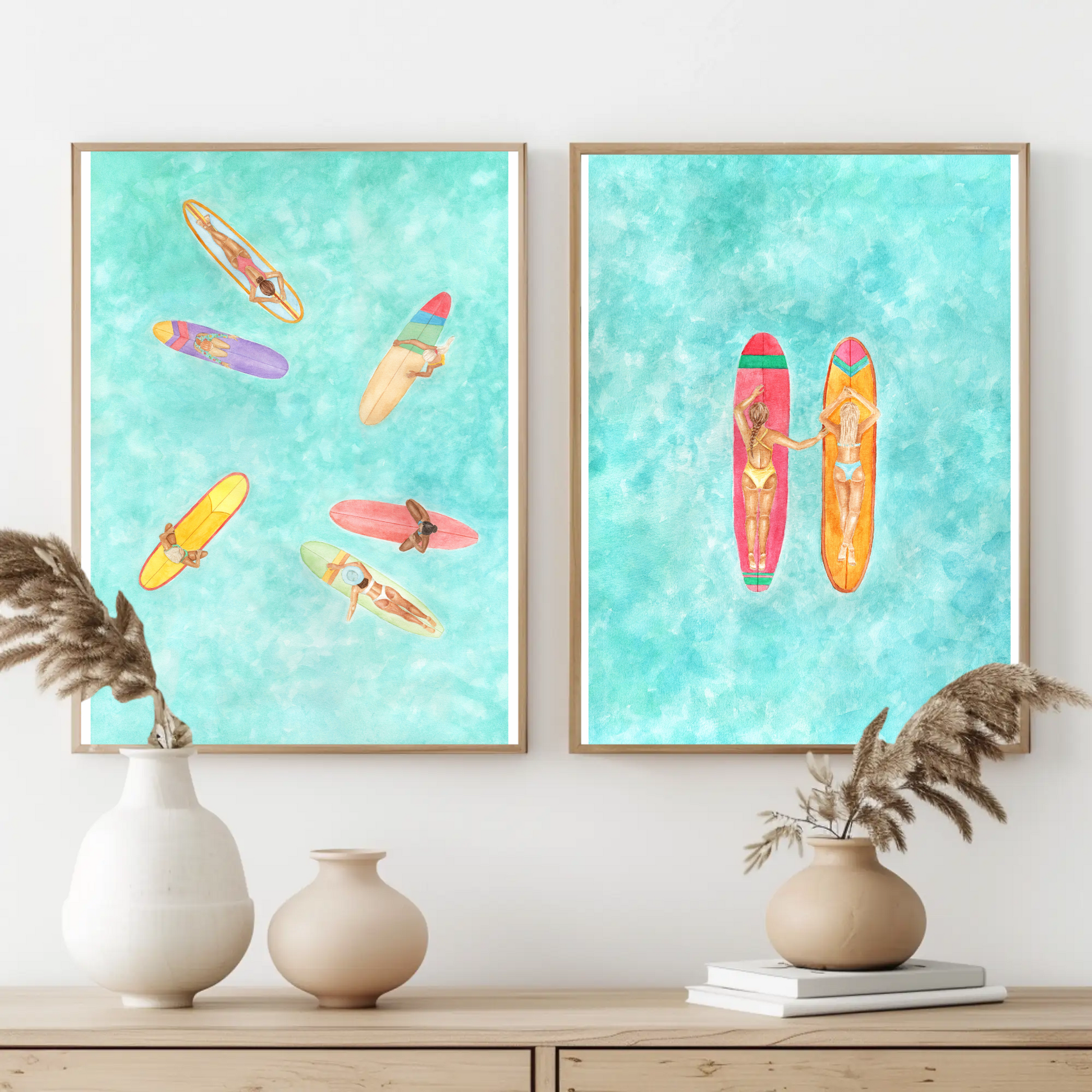 Two watercolour surf art prints with women surfers on longboards hanging on wall above shelf with vases and books in neutral tones