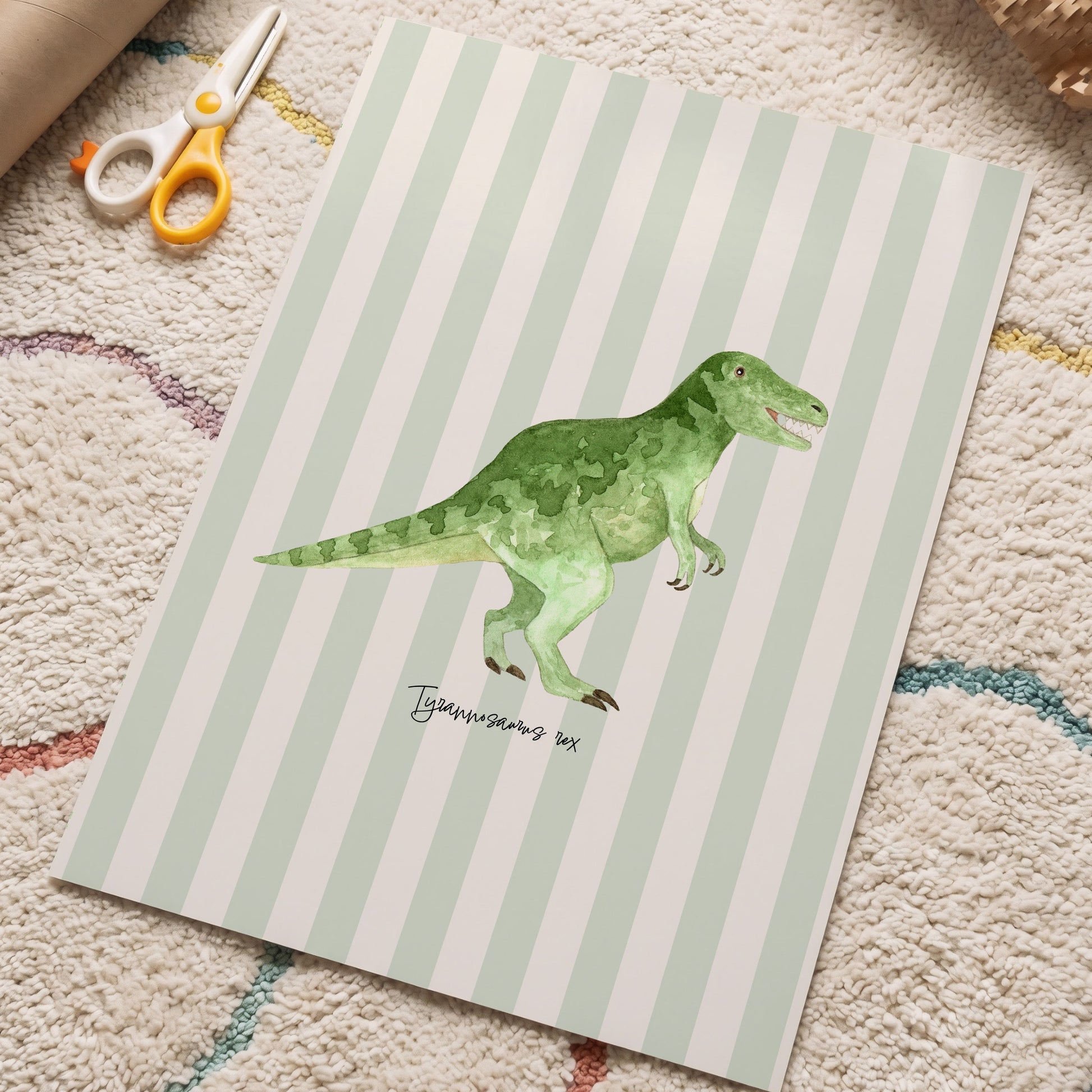 A children's art print featuring a green watercolor T-rex dinosaur on a mint green and white striped background, styled on a textured beige carpet with craft scissors and a cardboard tube nearby.
