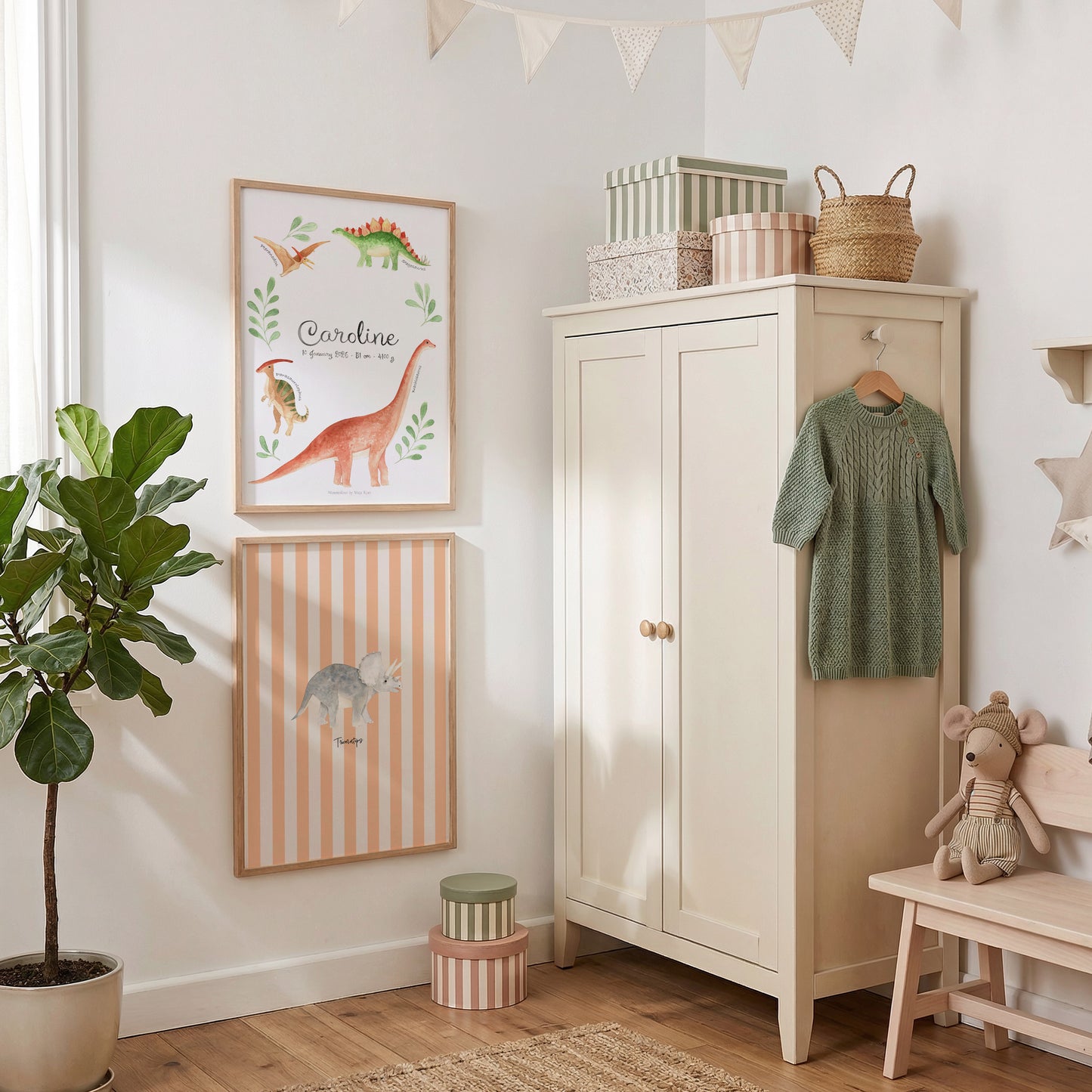 A styled children's room corner with two framed dinosaur-themed wall prints, a cream cabinet, a green knit sweater hanging on a hook, a potted plant, and a small bench with a stuffed toy.