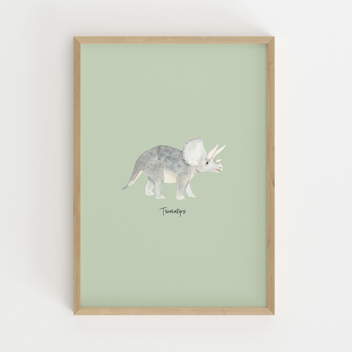 A framed art print featuring a watercolor illustration of a Triceratops dinosaur on a pale sage green background, enclosed in a light natural wood frame and mounted on a white wall.