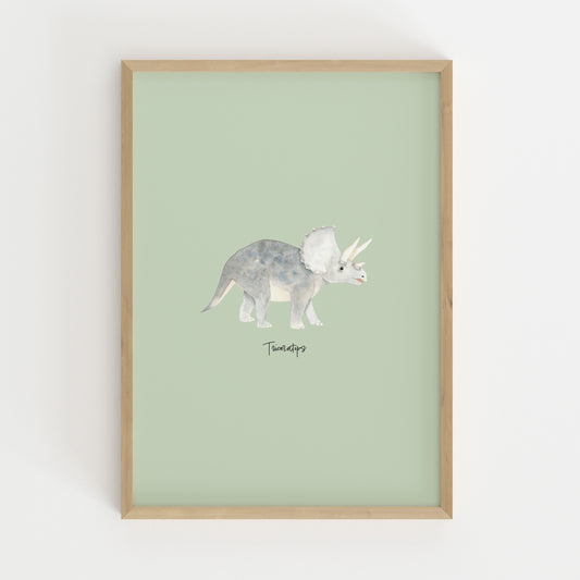 A framed art print featuring a watercolor illustration of a Triceratops dinosaur on a pale sage green background, enclosed in a light natural wood frame and mounted on a white wall.