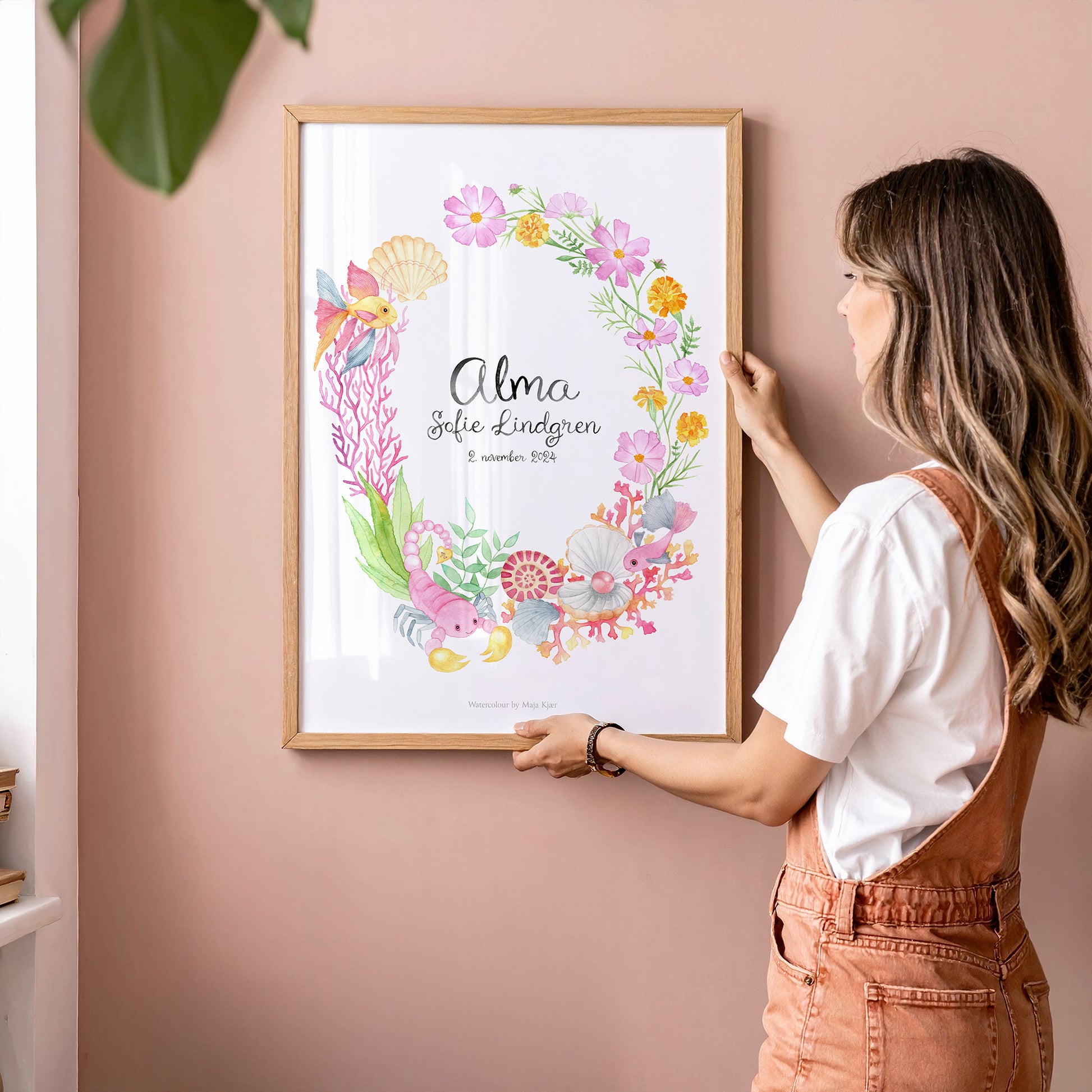 Woman hanging a framed floral wreath personalised baby name print with zodiac sign Scorpio on a pink wall.